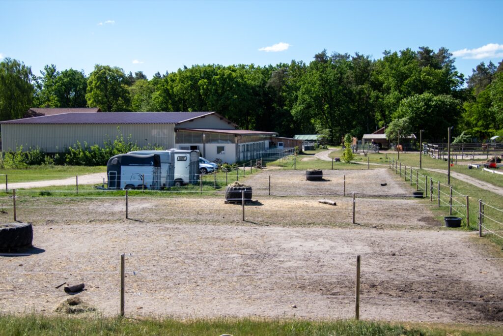 Pferdehof mit einem Reitplatz, Stallgebäude und einem Anhänger im Hintergrund.