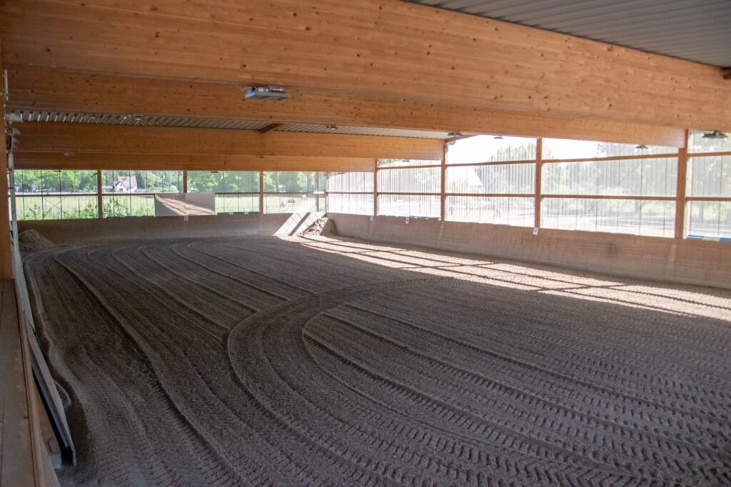 Reithalle mit einem sandigen Boden, der Hufabdrücke zeigt, in einem überdachten Bereich.