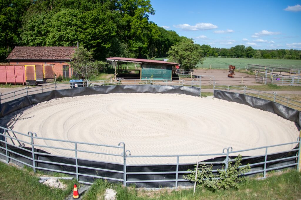 Runder Reitplatz mit Sandboden und Umzäunung in einer ländlichen Umgebung.