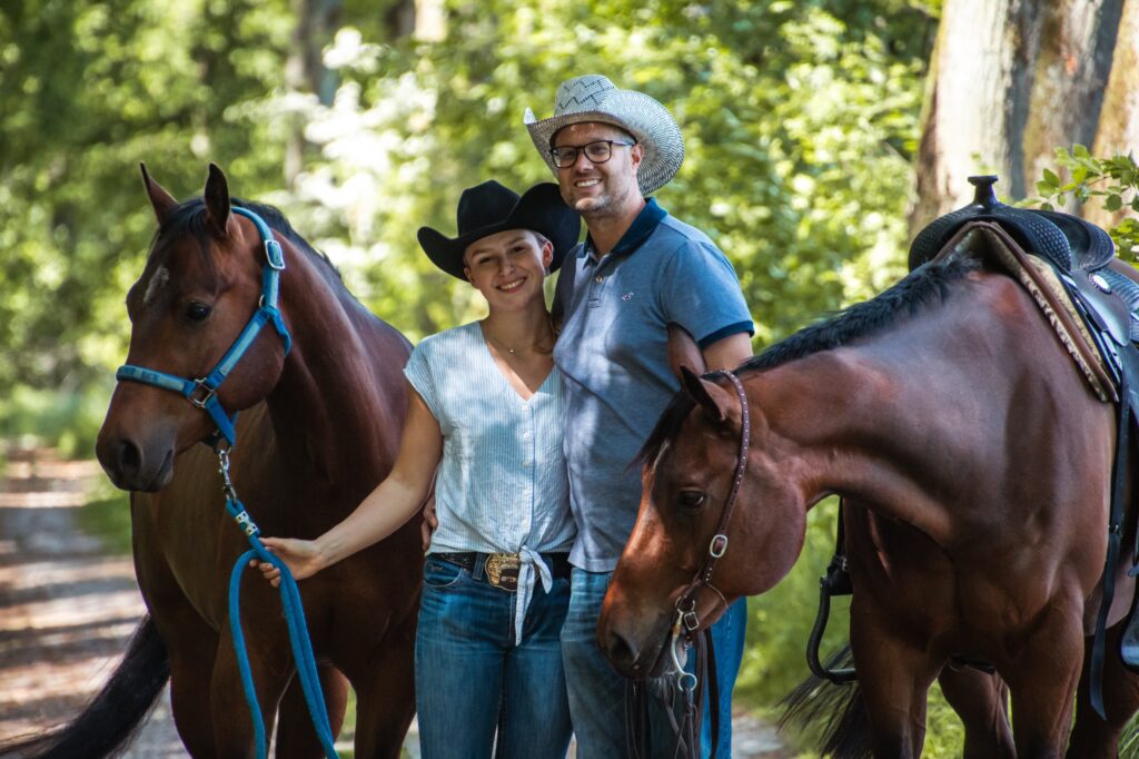 Ein Mann und eine Frau stehen mit zwei Pferden in einem Waldgebiet, beide tragen Cowboyhüte.