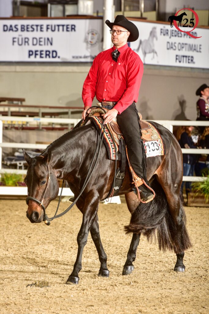 Mann im roten Hemd und Cowboyhut auf einem dunklen Pferd mit Westernsattel in einer Reithalle, Banner im Hintergrund, Szenerie eines Reitturniers.