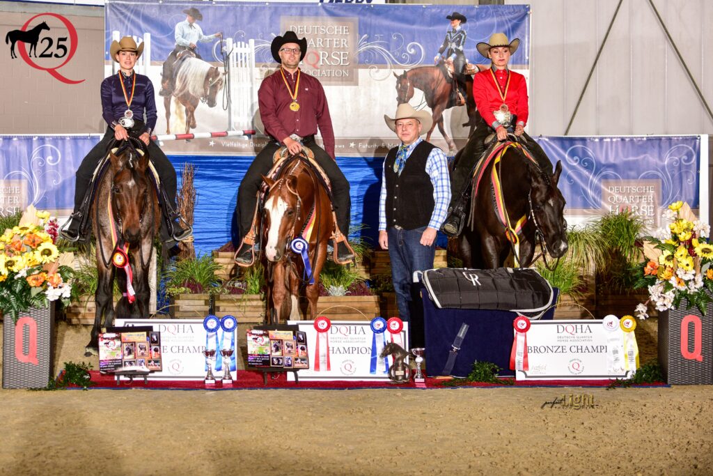 Vier Reiterinnen und Reiter auf Pferden stehen mit Medaillen auf einer Bühne vor einem blauen Banner, goldene Abzeichen, farbenfrohe Blumenarrangements und Pokale im Vordergrund.