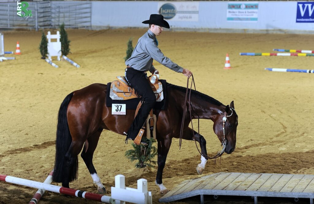 Reiter mit grauem Hemd und schwarzem Cowboy-Hut reitet ein braunes Pferd in einer Reithalle; Pferd trägt Leder-Sattel und Zügel, im Hintergrund Hindernisse und Werbetafeln.