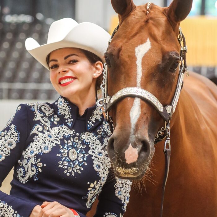 Amata Machner steht neben einem braunen Pferd mit heller Strähne, sie trägt einen weißen Cowboyhut und eine schwarze, reich verziert bestickte Jacke; Hintergrund einer Halle.