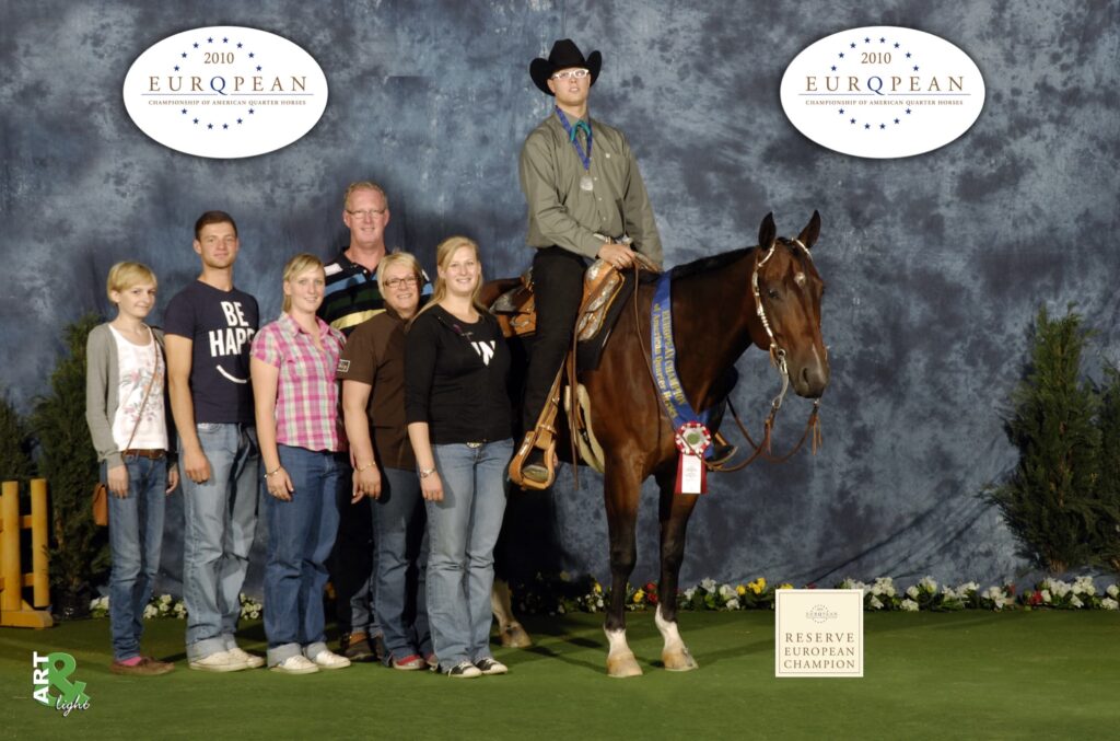 Gruppenfoto mit Reiter auf braunem Pferd, Medaillenkette, Siegerband und Hintergrund mit EU-Logo; Szene bei Alexander Eck Performance Horses, ruhig wirkende Besucherinnen und Besucher im Hintergrund