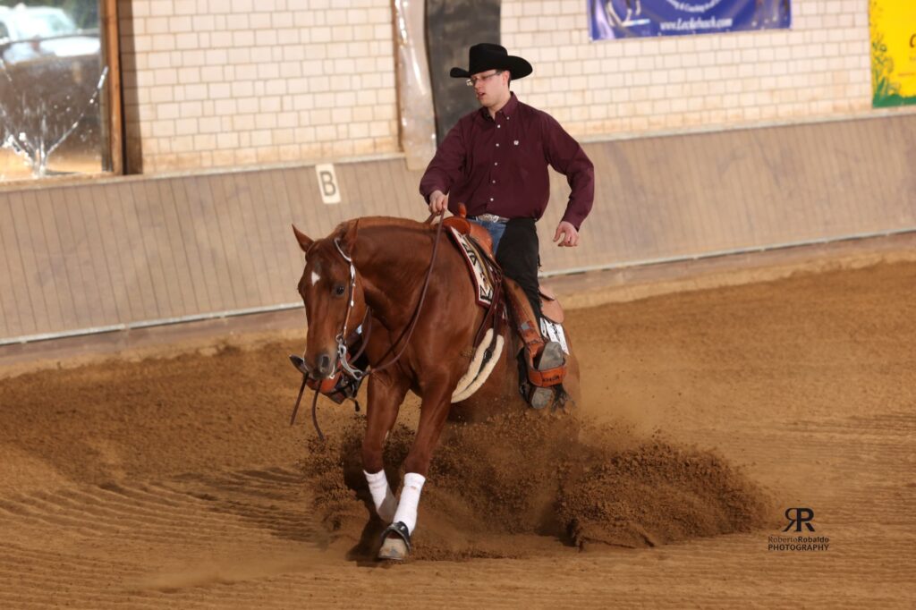 Reiter in braunem Hemd und Cowboyhut reitet ein braunes Pferd durch eine Sandarena in einer Indoor-Reithalle; Sand wird aufgewirbelt.