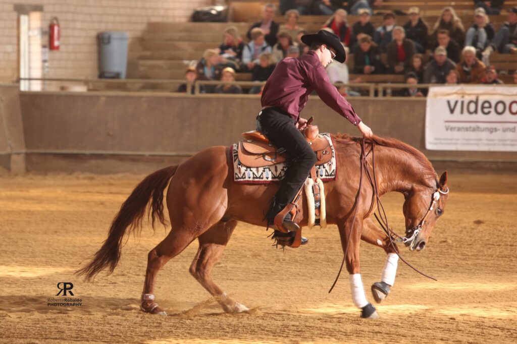 Reiter mit Cowboyhut und dunklem Hemd reitet auf braunem Pferd mit Westernsattel in einer Reithalle, Zuschauer im Hintergrund und Banner am Rand der Arena.