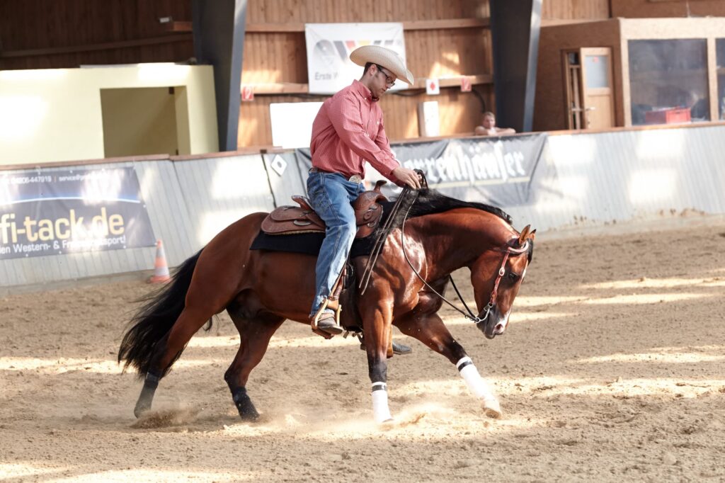 Reiter in rotem Hemd und Cowboyhut sitzt auf einem kastanienbraunen Pferd mit weißen Beinschonern in einer Reithalle, während eines Western-Trainings; Hintergrund mit Werbebannern und Zuschauern