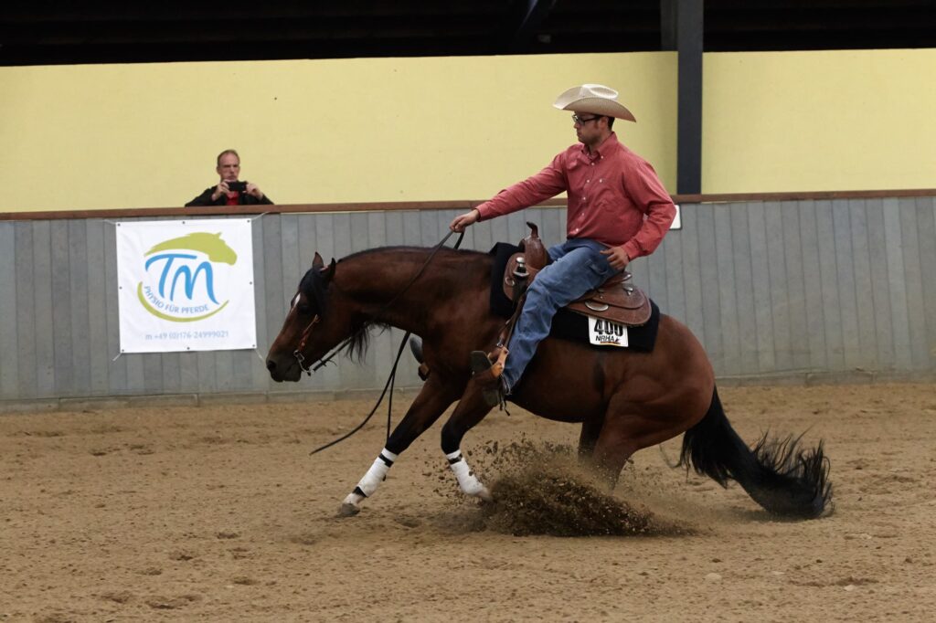 Reiter mit Strohhut und rotem Hemd reitet braunes Pferd in einer Indoor-Reitbahn, Staub wirbelt vom Hufschlag auf, Zuschauer fotografiert im Hintergrund.